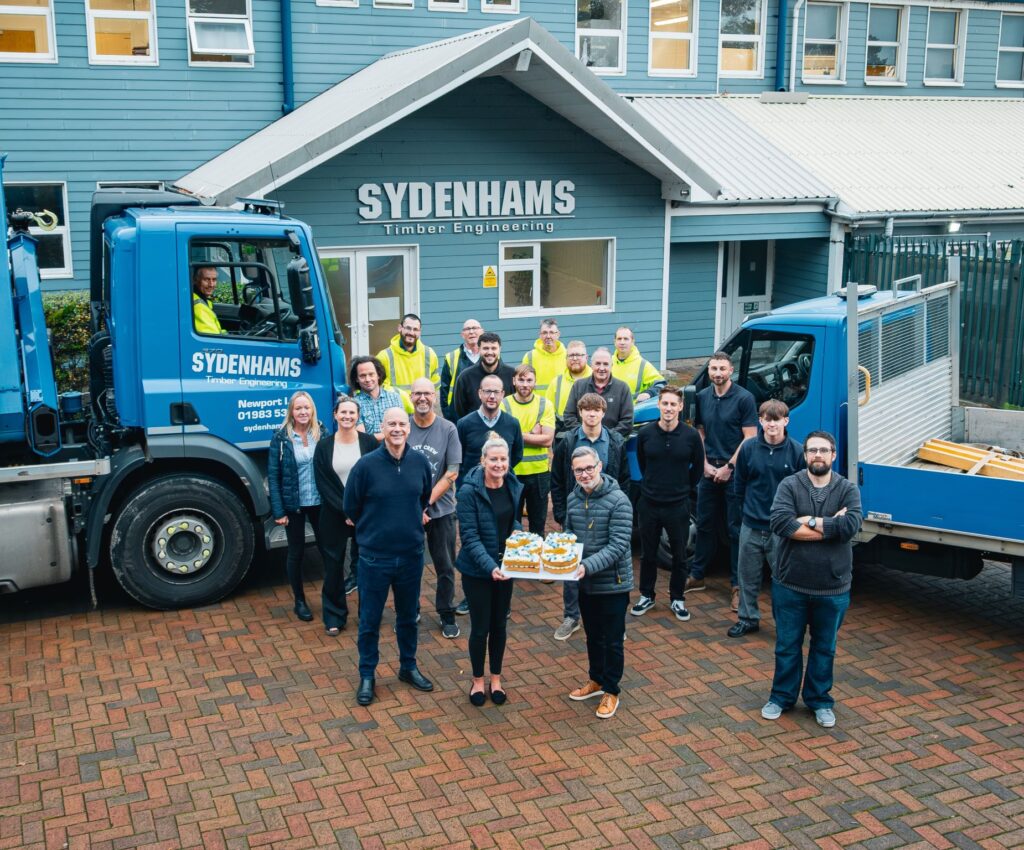 Employees celebrate Sydenhams Timber Engineering 25 years outside the Wickham timber factory with trucks and cake.
